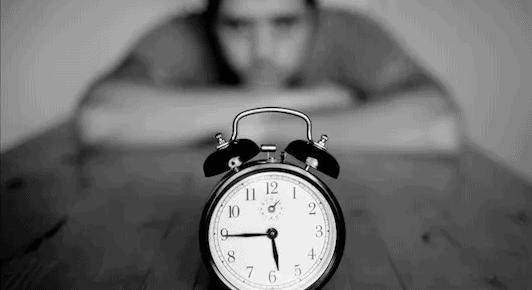 black and white image of man with chin on arms crossed on the table waiting and looking at a clock