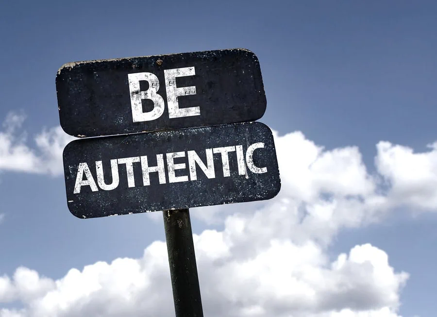Black sign against blue sky and puffy clouds saying Be Authentic